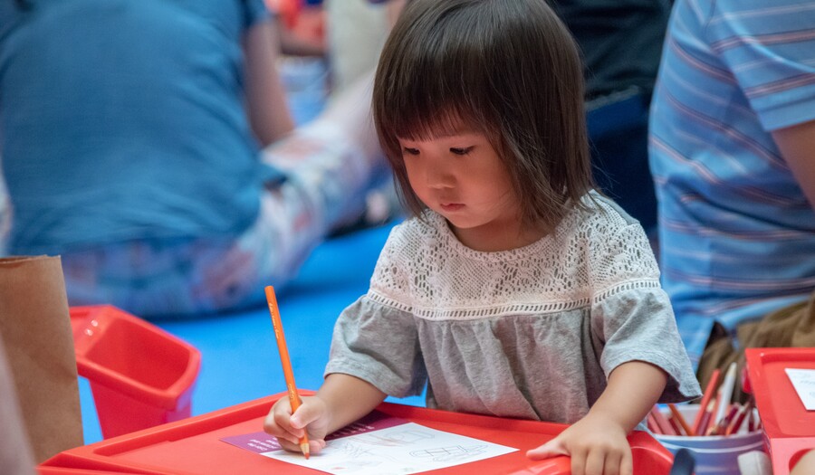 A toddler colors with a pencil at a red lap desk.