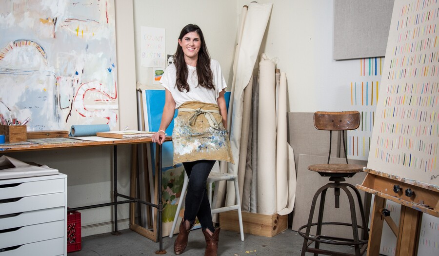 Artist Rachel Nash stands in her art studio surrounded by paintings and canvases.