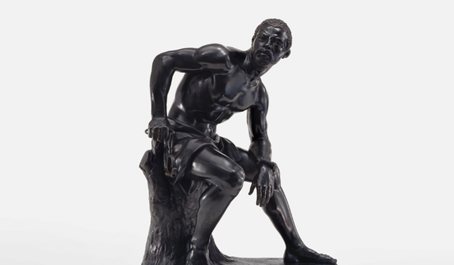 A bronze sculpture of a muscled Black man with manacles on his wrists leaning forward as he sits on a tree stump.