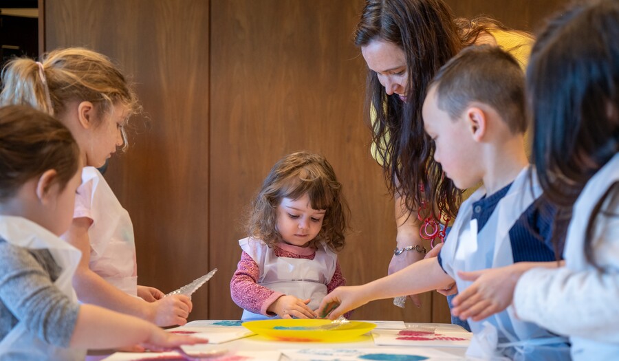 Older children and an adult help a toddler make art at a table.
