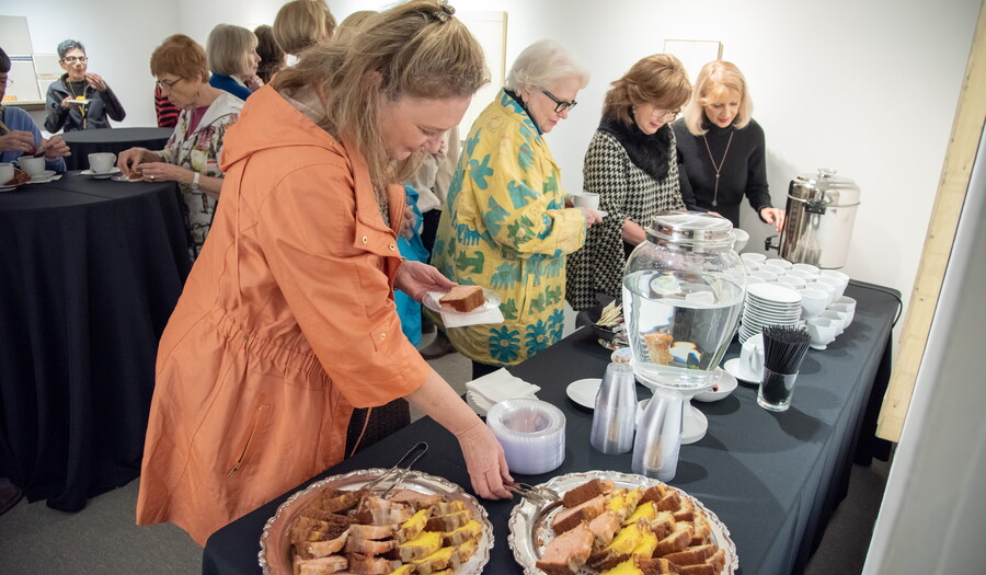 Adults stand in front of a table serving themselves tea and pastries.