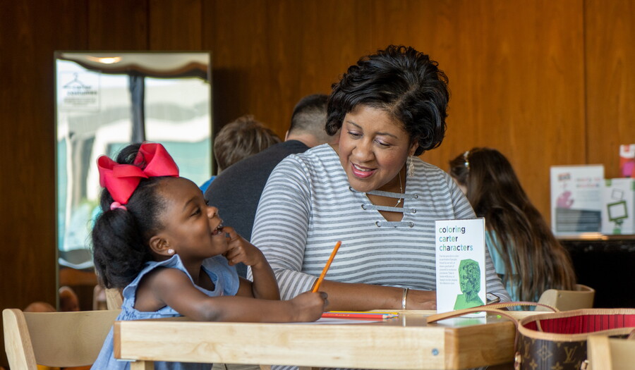 A toddler colors at a table while an adult watches.