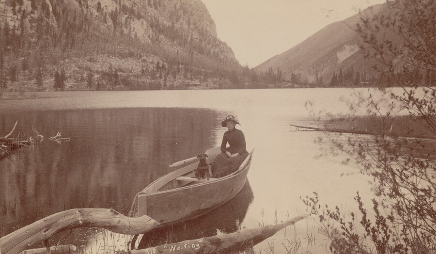 A sepia-toned photograph of a woman and her dog sitting in a canoe on a very still lake set in the mountains.