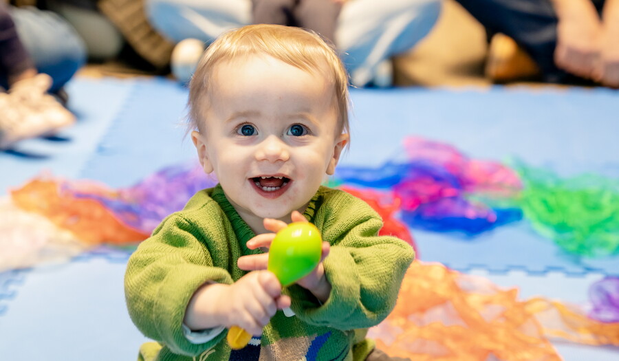 A smiling baby sits on the floor holding a green toy in their hands.