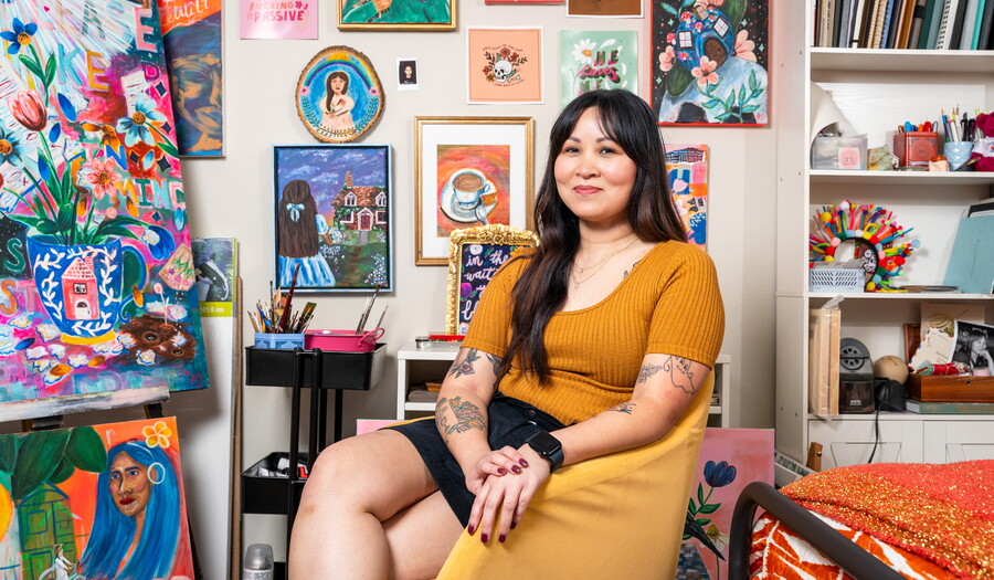 A smiling Asian woman with long dark hair is seated in a room with walls covered with colorful artwork.