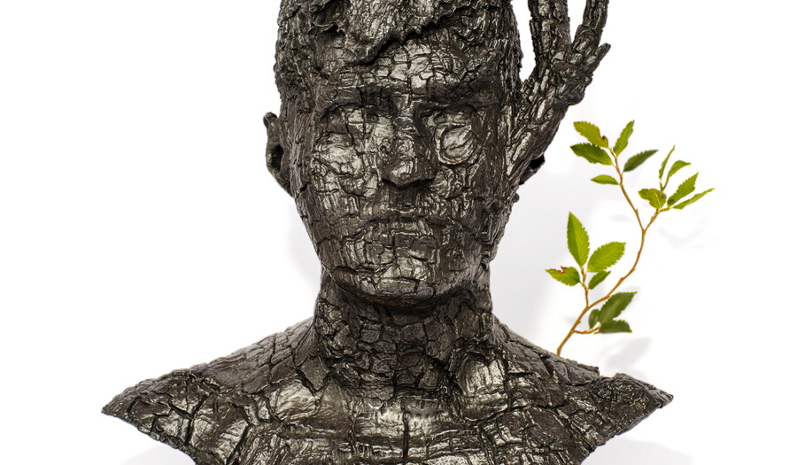 A bronze sculpture of the bust of a man made to look like it was carved from burned wood, with a sprig of green leaves sticking out from one shoulder.