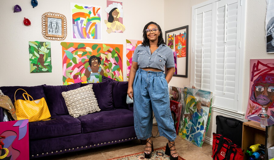 A smiling Black woman wearing glasses stands in front of a purple couch above which hangs many colorful artworks.