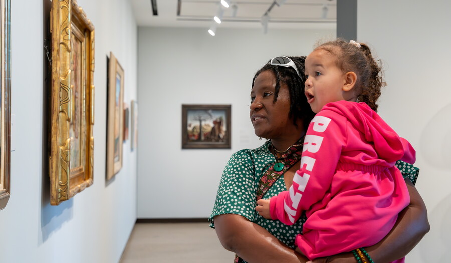 A Black woman holds a child in her arms as they look at artwork in a Carter gallery.