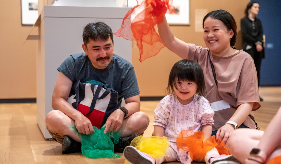 Two adults and a child sit on the floor of a Carter gallery playing with and waving colorful fabric.
