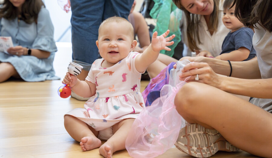 A White baby sits on the floor of a Carter gallery, adults nearby, waving and smiling.