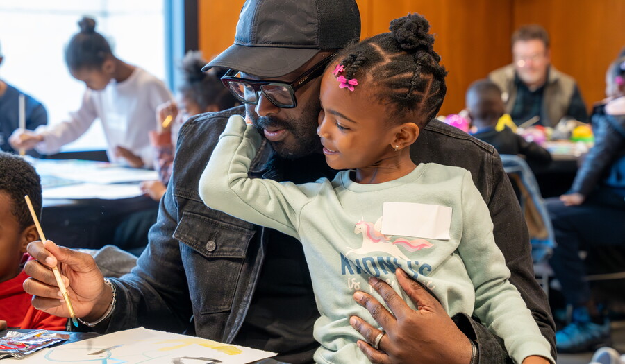A smiling Black child sits on the lap of a Black man as they paint together in the Carter&#039;s Pop-up Lounge.