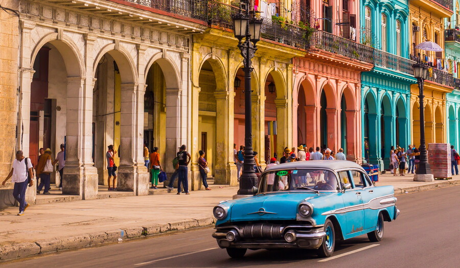 A street lined with colorful buildings and pedestrians walking along the sidewalk and under a street-level arcade; a 1950s-style blue car drives on the road.