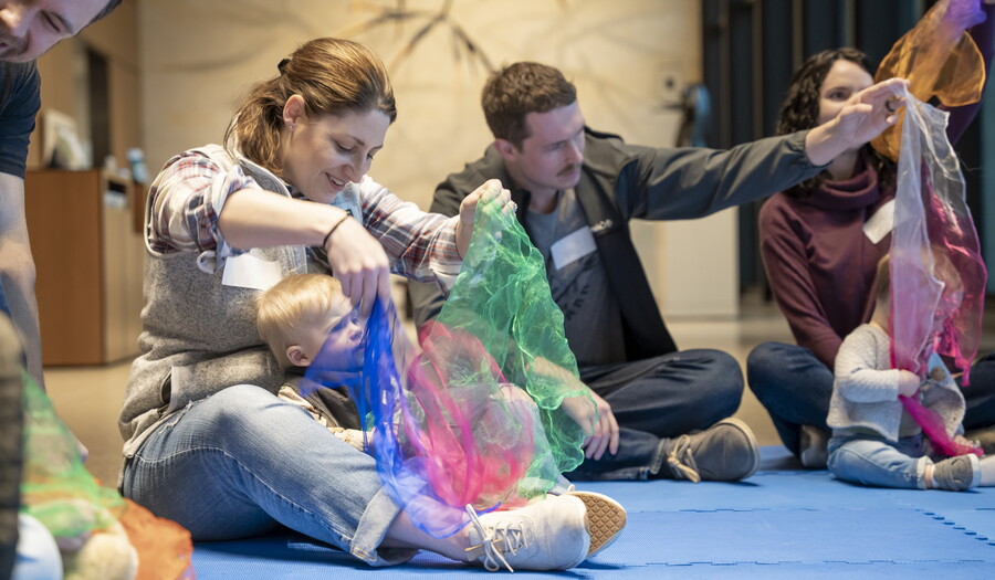 Several adults sit on the floor of a Carter gallery with their babies in their laps playing with colored cloths.