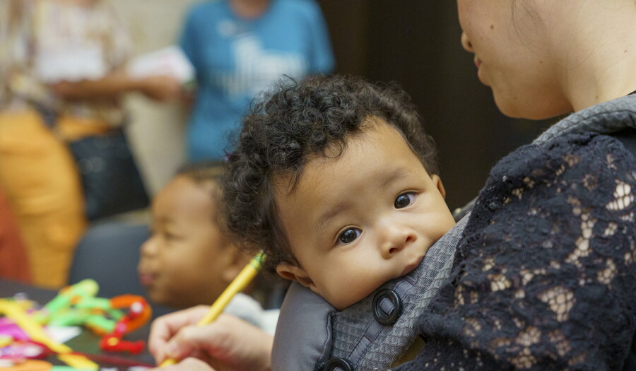 A baby with a full head of dark curly hair rests in a sling as the adult holding it makes art.