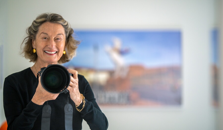 A smiling older White woman holds up a camera in front of her as if about to take a photo of the viewer.