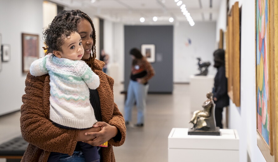 A Black woman holding a baby in her arms looks at a sculpture in a Carter gallery.