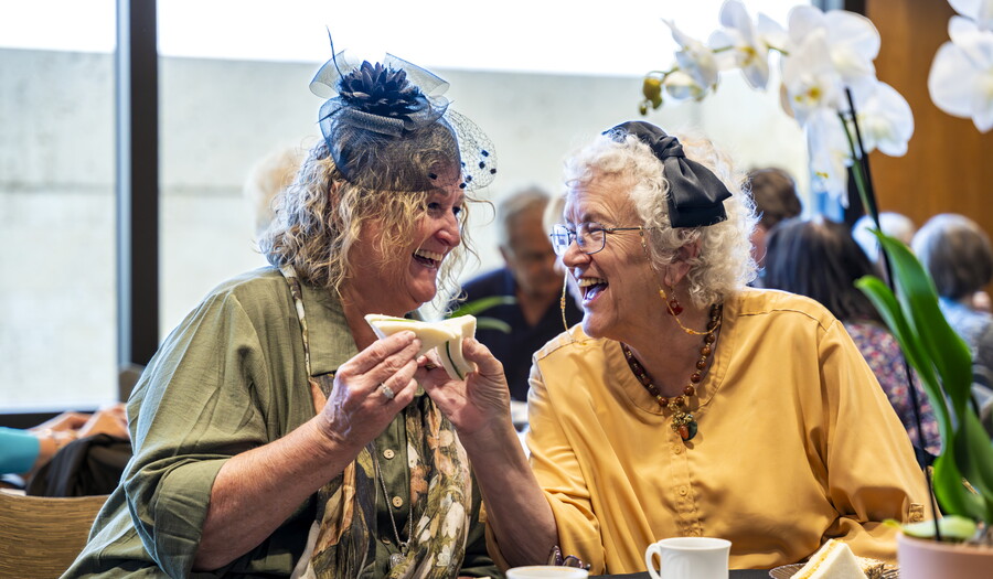Two older White women, both wearing hats, laugh together as they enjoy sandwiches.