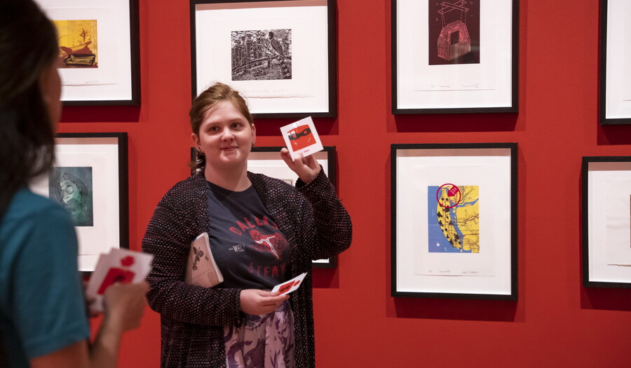A young White woman stands in front of artwork in a Carter gallery holding up flash cards for someone out of the frame.