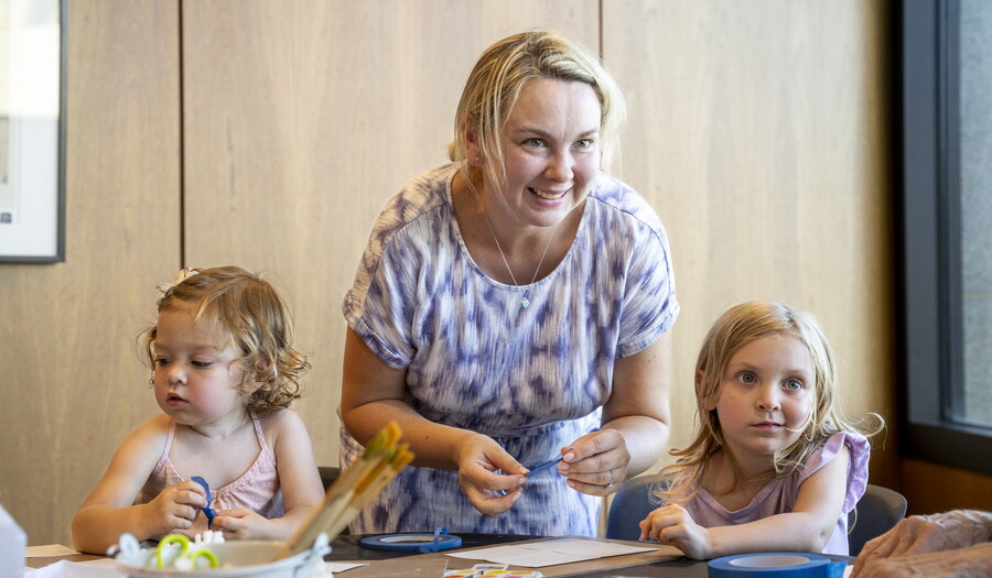 A White woman leans over a table in the Carter's Lounge where two children are making artwork.