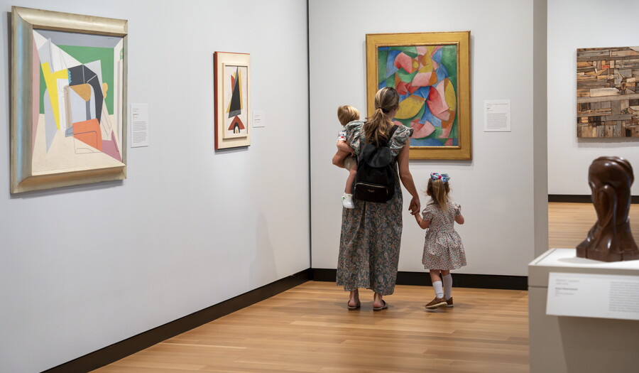 A woman and two children, one she holds on her hip and she hold the hand of the other, look at colorful art in a Carter gallery.