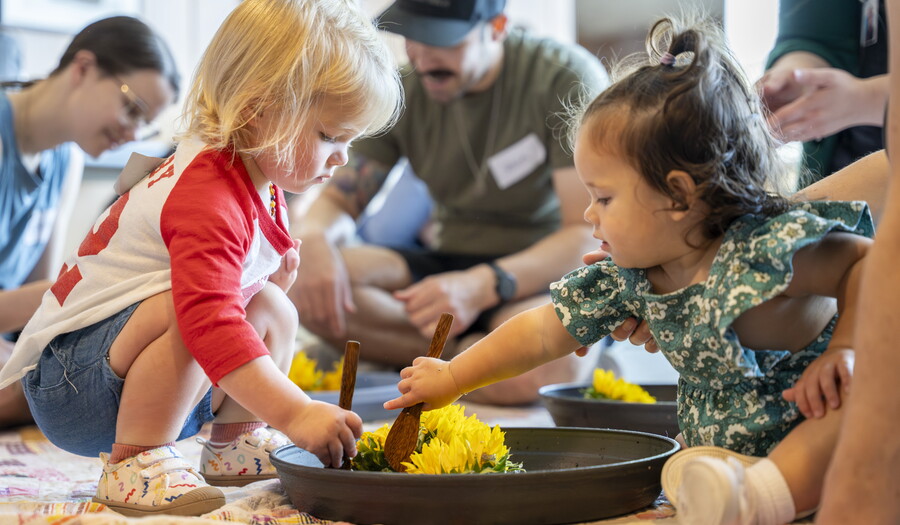 Two very young children play together with colored flowers in a bowl on the floor of a Carter gallery.