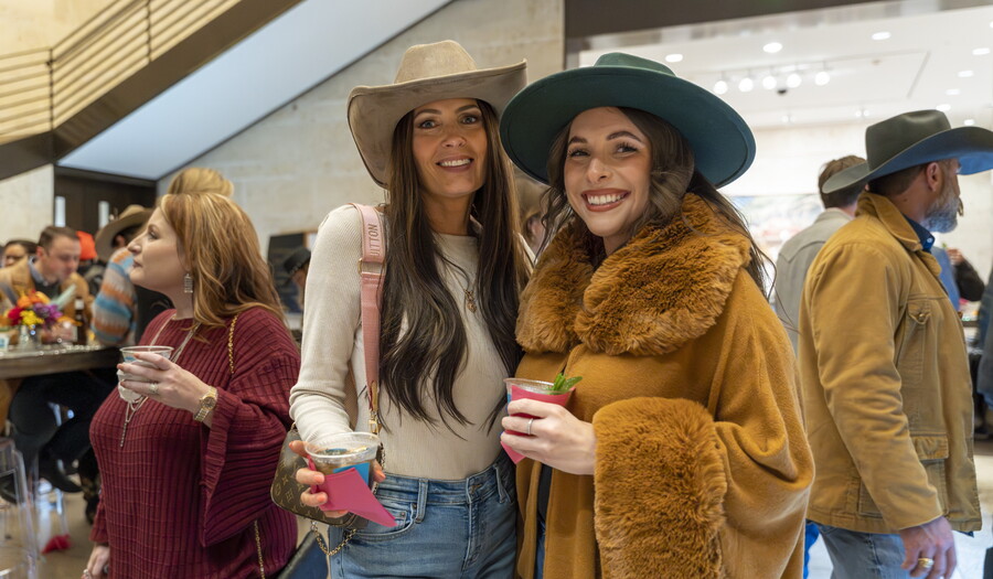 Two-light-skinned women stand side-by-side in the Carter's Atrium, both wearing cowboy hats and holding drinks.