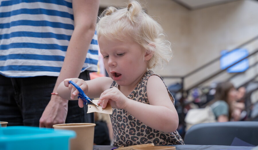 A White toddler uses safety scissors to cut paper while making an art project in the Carter's Atrium.
