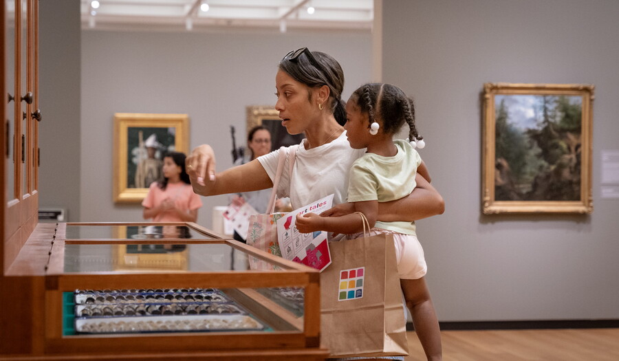 A dark-skinned woman holding a child on her hip points to some of the objects in "The Texas Cabinet."