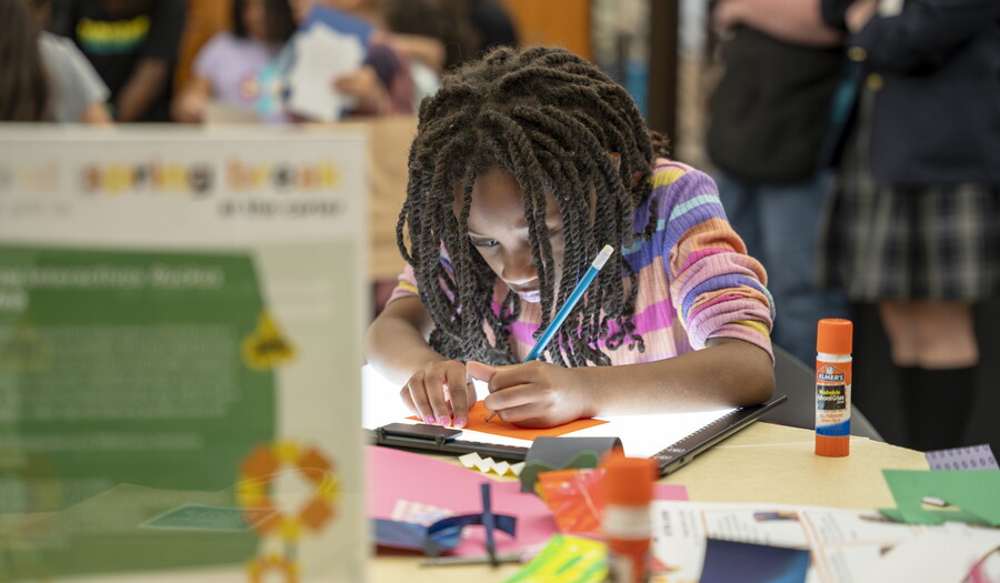 A black tween, focused on making art, leans over a table in the Carter's Library.