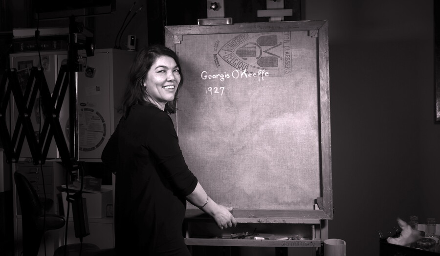 A black-and-white photo of a medium-skinned woman standing next to a canvas showing Georgia O'Keeffe's signature on the back.