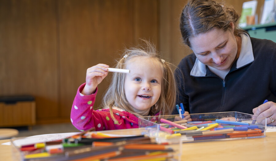 A young White child holds up a crayon as the adult next to her colors at a table in the Carter's Family Pop-Up Space.
