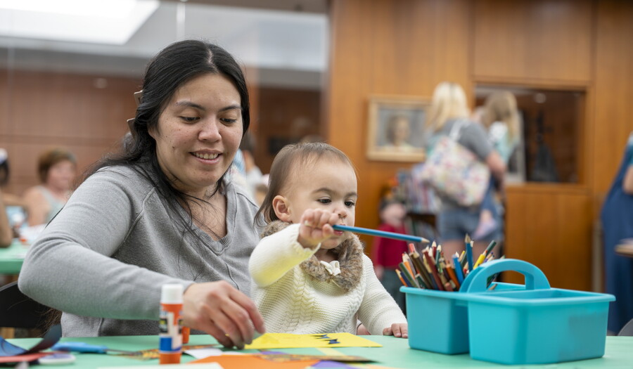 A medium-skinned woman with a baby on her lap sits at a table in the Carter's Library making art.