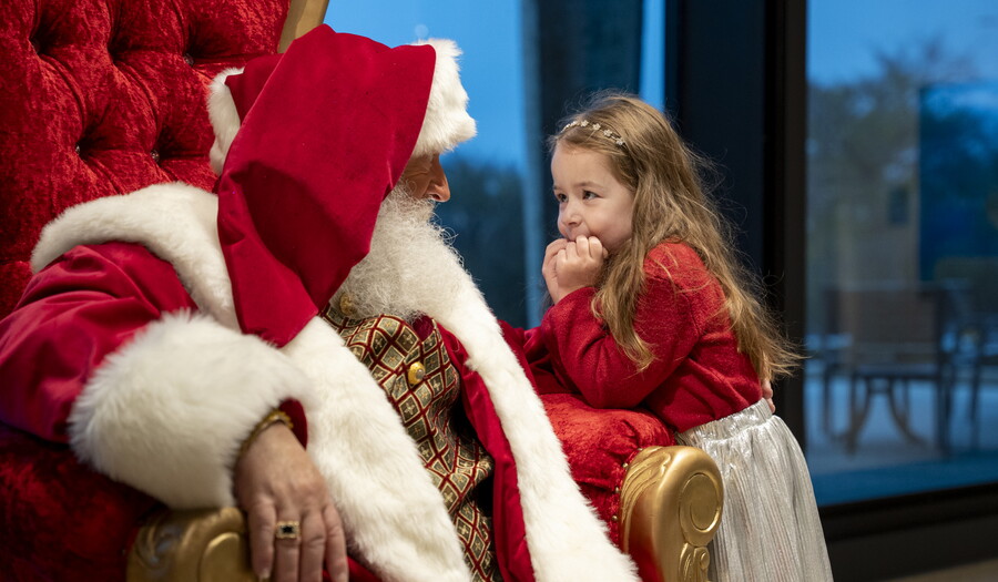 A young White girl leans on her elbow toward Santa who is seated in his throne.