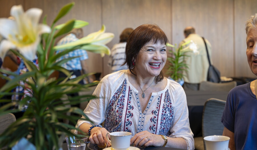 A smiling older White woman looks from behind a vase of flowers while seated at a table drinking tea.