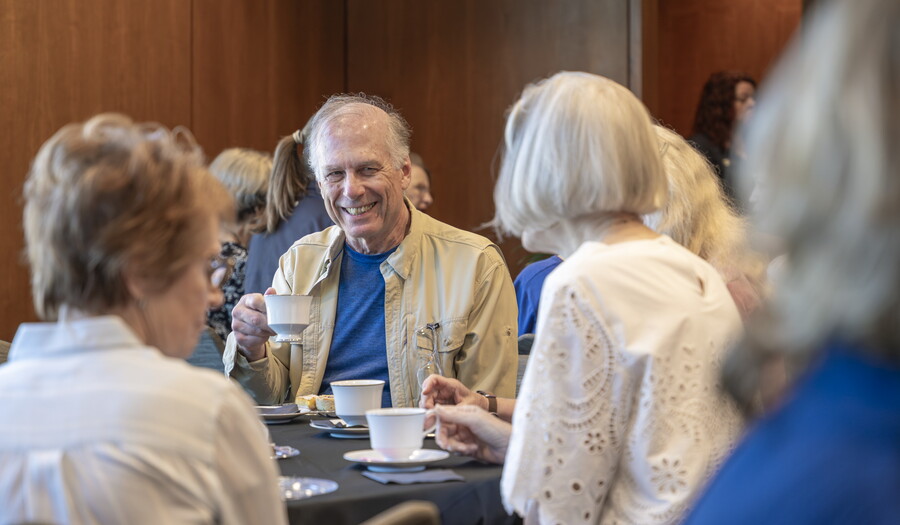 A smiling older White man drinks tea at a table with other guests.