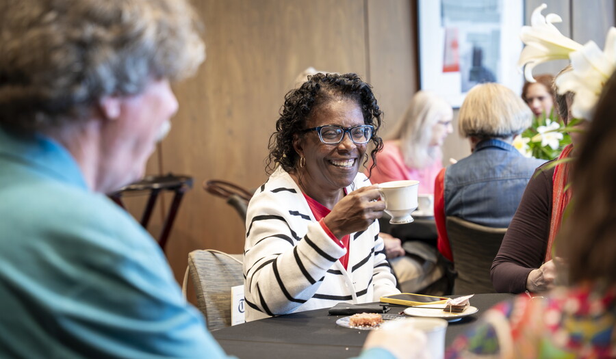 A smiling older Black woman holds up a cup of tea at a table in the Carter's Lounge.
