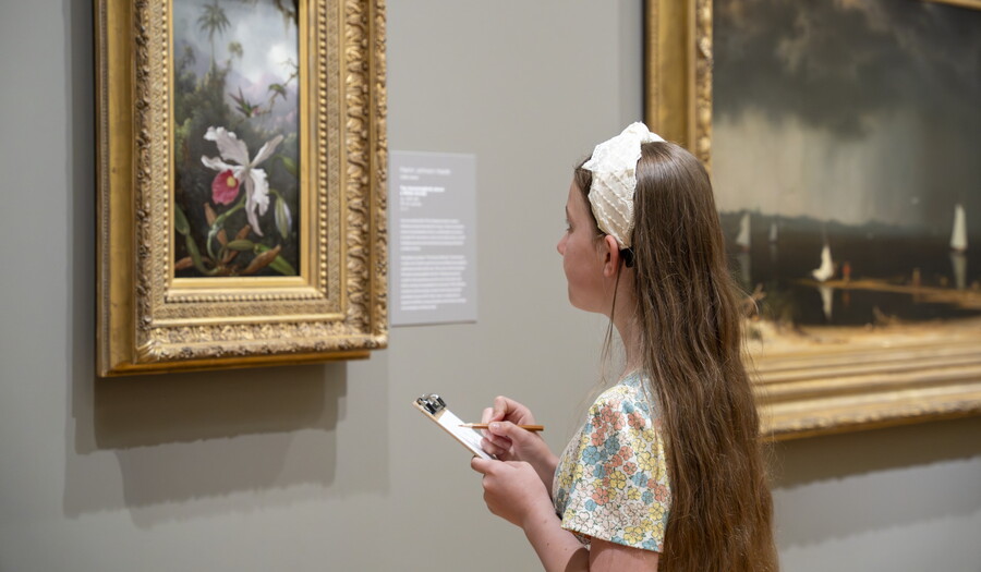 A White girl with long hair looks at an artwork in a Carter gallery and sketches it on a pad of paper.
