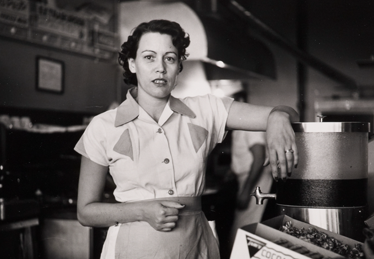 A black-and-white photograph of a White woman in a waitress uniform standing behind a diner counter with an arm propped on drink dispenser.