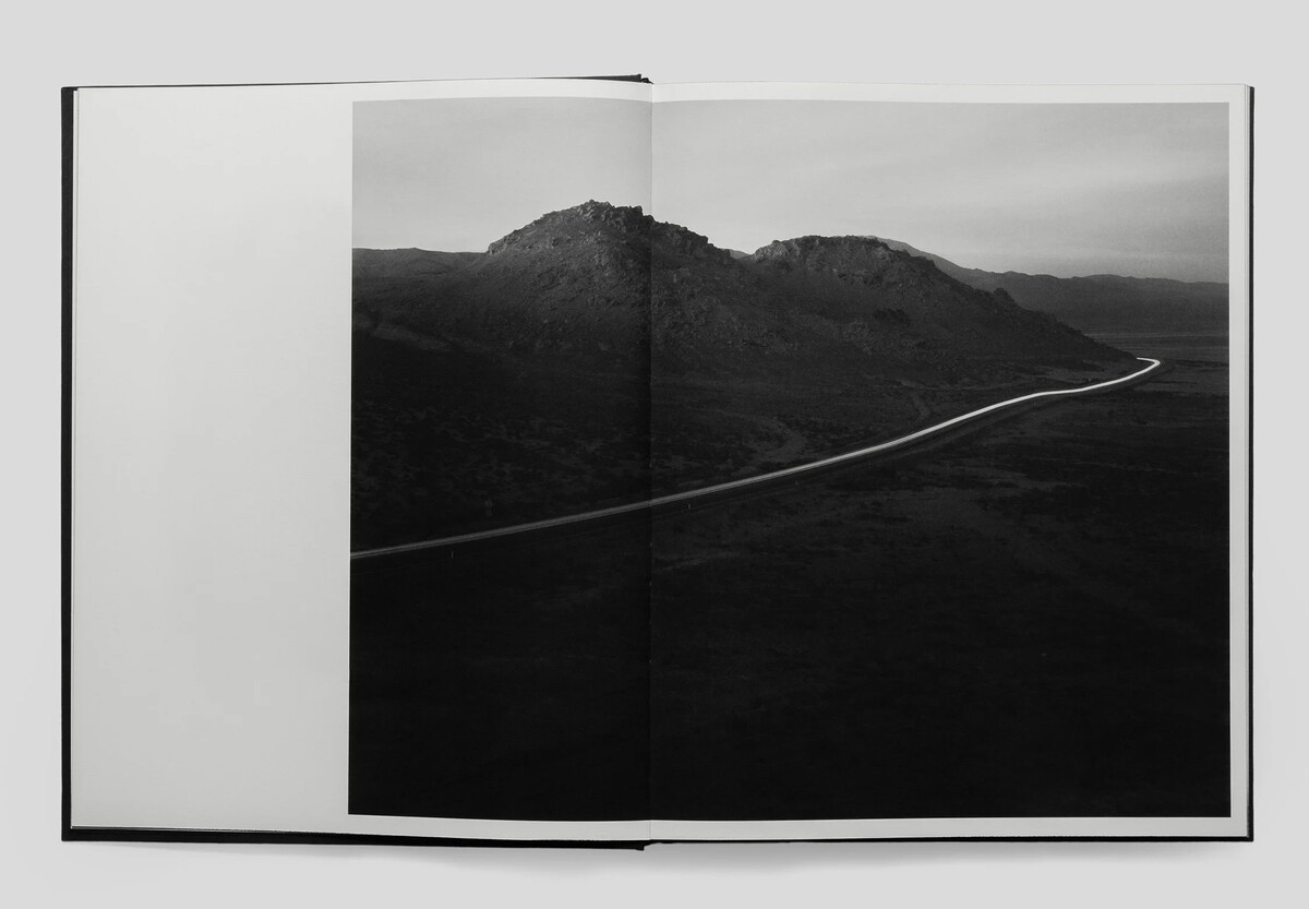 An open book with a black-and-white photograph spanning both pages of a lonely road bisecting the side of dark hills.