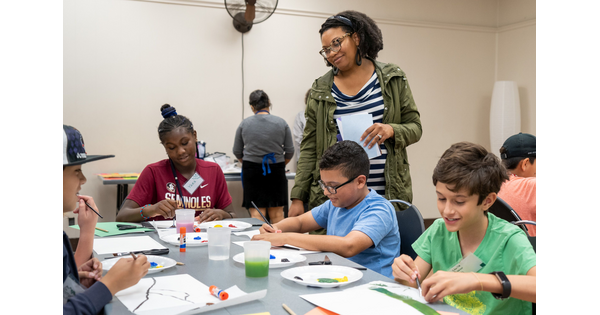 A standing woman looks at seated children doing an activity at a table.