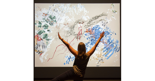 An adult make broad gestures in front of a screen with swaths of color on it.
