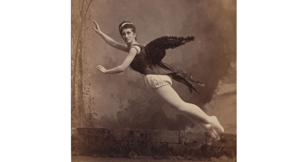 Black-and-white photograph from the 1880s of a woman wearing a dark corset with black feathered wings attached to the back and posed as if she is flying