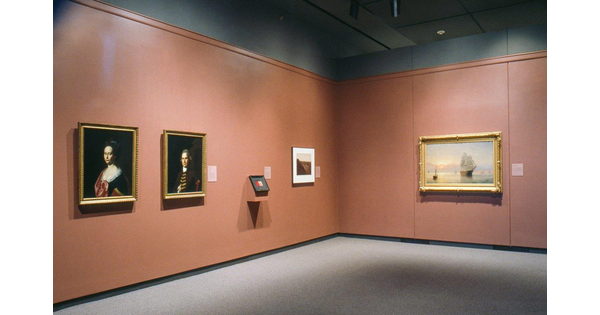 Artwork hangs on a taupe-colored wall. From left to right: a painting of a woman, a painting of a man, a landscape painting, and a painting of a boat on water on the far wall.