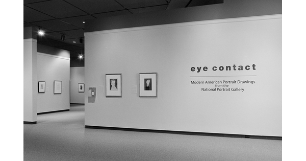 Black and white photo showing title wall of exhibition with two framed artworks to the left of the title. More artwork can be seen through the entryway.