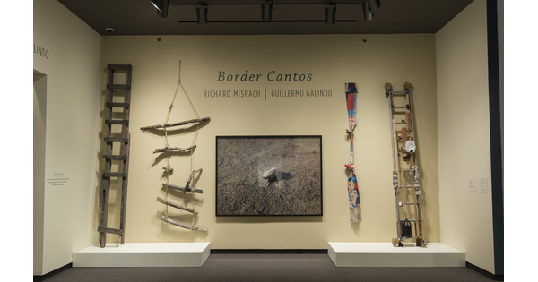 Cream-colored exhibition title wall with photograph of a rock in center. To the left are two crudely-made wooden ladders; on the far right is a ladder covered with detritus, and to its left is a knotted rope made from fabric.