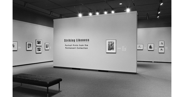 Black-and-white photo of gallery featuring the title wall with a framed portrait; in the background are more walls with framed artwork.