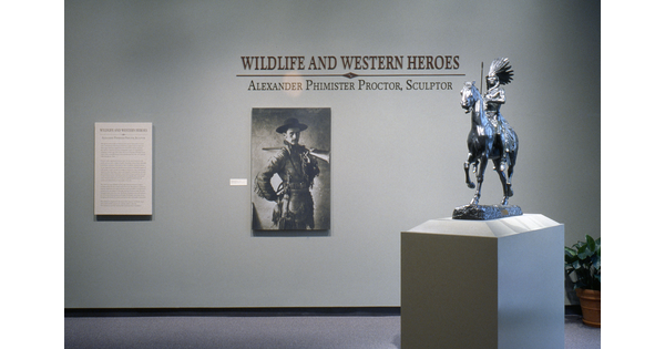 Under the title on a light gray wall is an enlarged photo of the artist dressed in cowboy garb; in the foreground on a pedestal is a sculpture of a Native American man riding a horse.