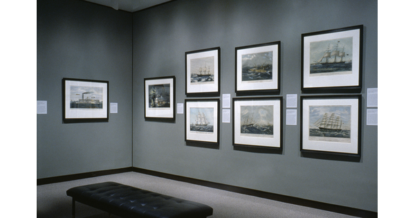 Corner of a gallery with gray walls; seven framed prints hang on the right wall, and one framed print hangs on the left wall.