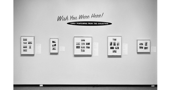 Black-and-white photo of a gallery wall showing five framed artworks, each featuring several postcards.