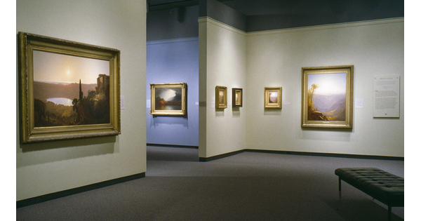 View of a gallery showing two large and three small framed paintings hanging on cream walls; through a doorway, another framed painting hangs on a blue wall.